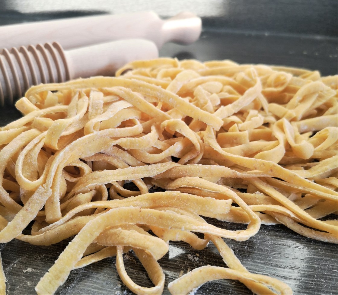 Eine Hand voll selbstgemachter, frischer Tagliatelle. Die Pasta ist bemehlt. Im Hintergrund sind die Nudelhölzer zu sehen.